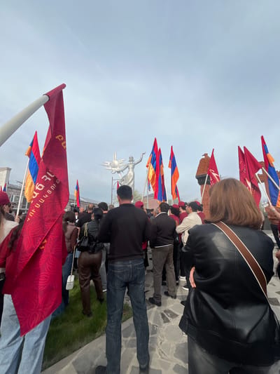 A crowd of people stand on a stone-paved area in Akhuryan, Armenia, with flags, some emblazoned with text, in the colors of the Armenian flag, red, blue, and orange. Some people are holding the flags. At the center of the view is a statue of a woman holding up a sun. The sky is overcast.