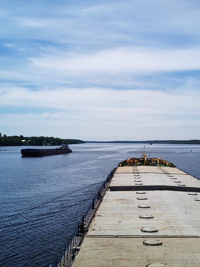 The image presents a wide-angle view from the aft end of a large, flat-decked barge, looking forward over a broad expanse of deep blue water. The sky occupies the upper third of the frame, displaying a light blue hue with scattered, wispy white cirrus and altocumulus clouds.

In the foreground, the concrete-colored deck of the viewing barge dominates the lower right, extending forward into the middle ground. The deck is marked by multiple parallel rows of uniform, circular, raised metal hatches. A blue railing system lines the port side of this barge, partially visible. Towards the bow, a compact superstructure with yellow and white components, possibly including a control cabin or machinery, is visible.

To the left of the viewing barge, another large, dark-hulled barge with a visible wake traverses the water, moving from left to right. This passing barge features a small, light-colored superstructure at its stern and appears to be carrying bulk cargo. Its movement creates clear ripples and a V-shaped wake pattern on the water.

The waterway extends to a distant horizon, bordered on both sides by continuous lines of dense green forest. Faint indications of small structures are discernible within the treeline on the left bank. In the far distance, approximately centered on the water, a small, dark vessel is visible as a silhouette, and two white navigational buoys are present in the middle-distance on the right side of the water.

The scene depicts daylight conditions consistent with a large river or lake. The image contains no specific geographic landmarks, signage, or urban features to definitively confirm the stated location as Mitino, Russia.