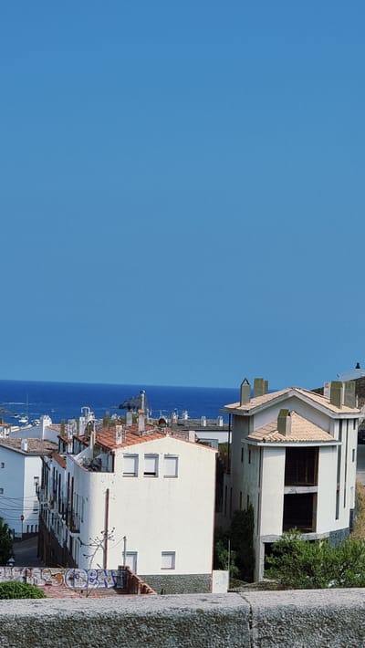 The image shows a view of the town of Cadaqués, Spain, from an elevated perspective. In the foreground, there is a stone wall with visible graffiti. Behind the wall, there are several white buildings with red-tiled roofs and numerous chimneys. The buildings are densely packed together, characteristic of a Mediterranean coastal town.
In the middle ground, a body of blue water, presumably the Mediterranean Sea, is visible. A small island with a structure resembling a lighthouse or a watchtower is situated in the sea. Several sailboats are dotted on the water, suggesting a harbor or anchorage area. The sky is clear and blue, indicating a sunny day. The overall scene depicts a coastal town in a sunny climate, with the sea playing a prominent role in the landscape.