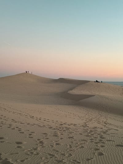 The image depicts a vast expanse of sand dunes, with the sky transitioning from a pale blue at the top to a soft pink hue towards the horizon. Footprints are clearly visible across the sandy surface, indicating recent human activity. Several small groups of people are scattered across the dunes. One group of approximately five individuals stands near the crest of a dune in the mid-ground, appearing as dark silhouettes against the lighter sand. Further to the right, another group of people is gathered on a higher dune overlooking the ocean, which is visible as a thin blue line on the horizon. The overall scene suggests a peaceful and natural environment, characteristic of the Dune of Pilat in La Teste-de-Buch, France.