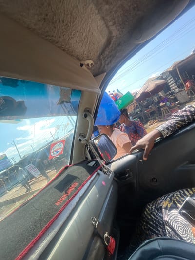 This media file captures a dynamic scene from inside a vehicle, likely a bus or van, looking out. The perspective is from the passenger side, facing forward and slightly upwards. The primary subjects are several individuals, with a focus on two women in the front of the vehicle. One woman, wearing a blue and red patterned top, is looking down, while another woman, in a light pink shirt, is looking towards the side. Their expressions are not clearly discernible.
The scene outside is a bustling market or roadside setting in Mowe, Nigeria. It appears to be daytime, with bright sunlight indicating a clear sky. The environment is filled with stalls, structures, and a crowd of people, suggesting a busy commercial area. There are various objects visible, including a large blue plastic bag, a green crate, and several bottles of what appear to be beverages, possibly soda. The vehicle's interior is also visible, with a textured tan panel on the left and a black dashboard area on the right. A grey suitcase with red trim is placed on the right side of the vehicle's interior.
A notable detail is a red flag with text that reads "CHAMPION" and "MUMBAI" with a circular emblem. There's also a red and black mat on the dashboard with the word "HIGHLANDER" in red lettering. The overall impression is one of vibrant activity and movement, characteristic of a busy market or transportation hub in Nigeria. The image appears to be taken during the day, with strong sunlight casting shadows and illuminating the scene.