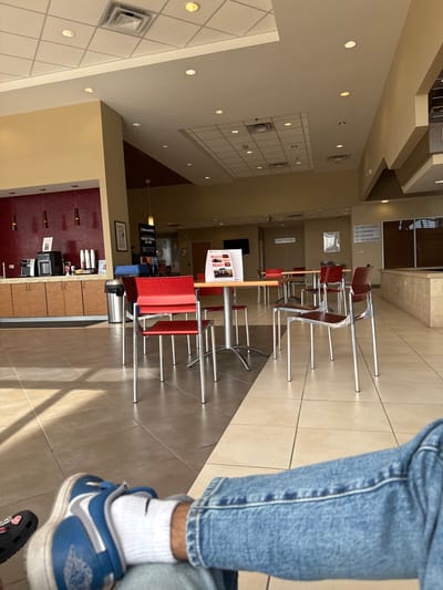 Here is a detailed description of the image:
**Scene:** The image captures the interior of a waiting area, likely within a business like a car dealership or service center. The space is well-lit with overhead lighting arranged in a grid pattern on the ceiling. The color palette is predominantly neutral tones—beige walls, tiled floors, and silver accents.
**Objects:**
* Rows of modern chairs and tables are arranged in the center of the room, providing seating for waiting customers. The chairs feature red seats and silver frames.
* A refreshment area is visible in the background, with counters, cabinets, coffee makers, and possibly other beverage-related equipment. The walls behind the refreshment area are decorated with red tiles.
* Signage on the walls includes directional information for restrooms, cashier, service, and retail parts.
* There is a display board featuring advertisements or informational material about "Road Trip Ready" and SiriusXM radio.
**People:**
* A person is partially visible in the foreground, wearing blue jeans, white socks, and blue Nike Air Jordan sneakers. Only the legs and feet are in view.
**Environment:**
* The space feels clean and organized, intended to provide a comfortable waiting experience.
* The mix of seating arrangements and amenities suggests that the area caters to both short-term and extended waiting periods.
**Location:** This image likely comes from the city of Palatine in the United States.