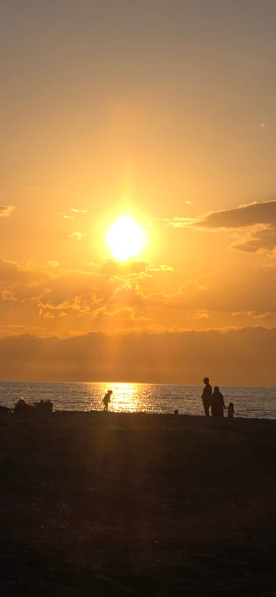 The image depicts a sunset over the Issyk-Kul lake near the village of Boz Beshik, Kyrgyzstan. The sun is a bright, intense orb in the sky, casting a warm, orange glow across the scene and creating a shimmering reflection on the water. The sky is filled with scattered clouds, some of which are illuminated by the setting sun, giving them a golden hue.
In the foreground, a silhouette of a beach or shore is visible, with a few figures of people scattered across it. To the left, near the water's edge, there appear to be some objects or debris. In the middle ground, a solitary figure, possibly a child, is silhouetted against the bright reflection of the sun on the water, bent over as if playing. Further to the right, another group of silhouettes, appearing to be a family with at least two adults and a child, stand facing the water and the sunset. The overall atmosphere is serene and tranquil, capturing the beauty of a sunset over a large body of water.