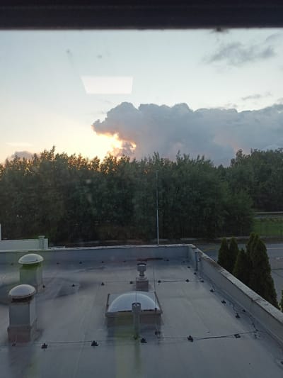 The image depicts a view from a window overlooking a flat rooftop and a treeline under a cloudy sky with visible sunlight breaking through. The rooftop features several ventilation domes, pipes, and wiring. Beyond the rooftop, a dense line of trees forms the horizon, with hints of sunlight illuminating them from behind. A street and some small evergreen trees are visible to the right of the treeline. The scene appears to be an outdoor environment in Biały Bór Drugi, Poland, during either sunrise or sunset due to the lighting conditions and cloud formations. No people or specific actions are discernible in the image.