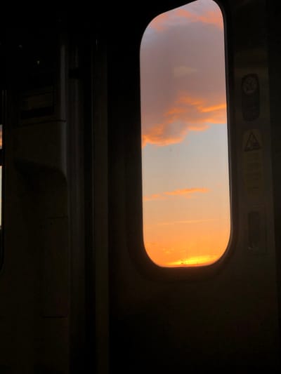 A view from inside a dark vehicle, possibly a train or bus, looking up through a rectangular window with rounded corners. Outside the window, the sky is a mix of orange and peach hues, likely during sunrise or sunset. Clouds with orange highlights are visible. The interior of the vehicle is dark, with the window frame and surrounding ceiling area in shadow.