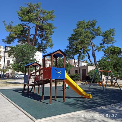 This is a full, eye-level shot of a children's playground in Ruvo di Puglia, Italy, on a sunny day. The playground features a wooden play structure with a blue roof, red and blue panels, and a yellow slide. To the right of the play structure, there is a swing set with two swings. The playground is situated on a dark green rubberized surface, bordered by a concrete path. In the background, there are large pine trees, lush green bushes, and buildings, suggesting an urban or suburban setting. A man wearing a black t-shirt and green shorts is standing near the swing set, accompanied by two dogs. Cars are parked along the street in the distance. The date and time stamp "06.07.2026 09:37" is visible in the lower right corner.