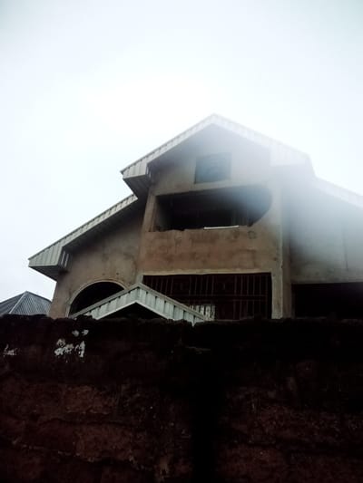 An overcast, hazy day in Nanka, Nigeria, showcases the unfinished construction of a multi-story building. The structure, primarily composed of concrete and featuring corrugated metal roofing, rises above a dark, rough-textured wall in the foreground. Architectural elements include a prominent arched opening and several windows, some with metal bars. The building appears to be in a state of disrepair or ongoing construction, with exposed concrete and an unpainted facade. The surrounding environment is obscured by the heavy fog, with only hints of other structures visible in the distance. The scene conveys a sense of incompletion and damp weather conditions.