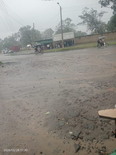 The image captures an outdoor scene on a wet, unpaved road in Kapkures ward, Kenya, at 17:30 on February 28, 2026. The environment is overcast, and the ground, comprising the foreground and midground, consists of mud and degraded asphalt with numerous potholes and standing water, consistent with recent precipitation. Scattered debris and litter, including a green wrapper, are visible on the ground. A partial wooden surface is visible in the bottom right corner.
In the midground, a group of approximately 15-20 individuals stands under a shelter structure along the right side of the road. Some individuals within the group are holding umbrellas. To their left, a green-roofed structure with partial lettering "O ASIA FOODS & BEVERAGES FARM" is present. A light-colored wall borders the road behind the group.
Two motorcycles, both equipped with umbrellas, are visible on the road. One, carrying a driver and a passenger, is positioned in the left-midground. A second motorcycle with an umbrella and at least one passenger is in the right-midground. A tall utility pole with multiple overhead power lines stands prominently on the left side of the midground.
In the background, vehicles including a red container truck, a white truck, and a dark-colored sedan are discernible. Trees line the distant horizon under an overcast sky.
