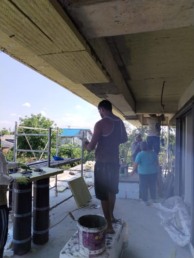 The image depicts an outdoor construction scene, likely in Dumbrăveni, Romania, with several individuals engaged in building or renovation work. The primary focus is on a group of people working on what appears to be the exterior of a building, possibly installing insulation or some form of exterior cladding on an overhang or balcony.

In the foreground, a man in a brown tank top and dark shorts stands on a white bag, holding a trowel and what seems to be a piece of insulation material. Another person in a white shirt and green gloves is seen to the left, also working with insulation and a trowel on a table supported by two dark, cylindrical objects wrapped in black material and silver tape.

Further back, two individuals are visible. One, wearing a light-colored shirt and blue jeans, is standing on a ladder and appears to be working on the ceiling structure, where insulation panels are being attached. Another person in a blue shirt and dark pants is standing nearby. The background reveals a residential area with houses, some with blue roofs, and lush green trees under a clear blue sky with scattered white clouds. The overall impression is a sunny day, consistent with construction work in pleasant weather.

Notable details include the scaffolding visible behind the table in the foreground, suggesting a multi-level construction project. The insulation material is light yellow and appears to be rigid panels. There are several buckets and construction debris scattered around the work area. Some visible text includes a label on the bucket in the foreground, partially obscured, and what might be a brand name on the tape wrapped around the cylindrical supports. The atmosphere seems to be one of active construction and labor.