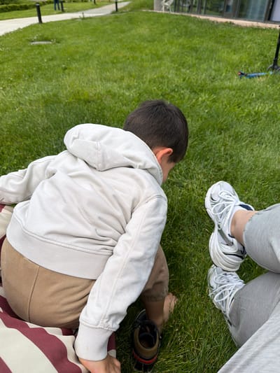 A child in a light-colored hooded sweatshirt and brown pants sits on a striped blanket on a grassy surface, presumably in Chkalov, Armenia. An adult wearing gray pants and white sneakers is partially visible in the top right corner of the image. The child has dark hair. There is footwear near the child. The background consists of green grass and a path with lampposts.