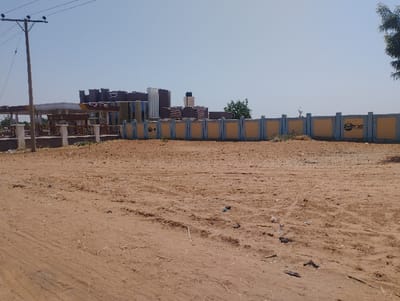 The image shows a large, empty dirt lot in Sangaro, Nigeria. The lot is located in front of a concrete wall with a blue stripe and a sign that reads "First Bank". The wall is approximately 10 feet tall and is located in front of a building. The building is a three-story structure with a flat roof and appears to be made of concrete. There are no people or vehicles visible in the image. The environment is dry and dusty, with minimal vegetation. The sky is clear and blue.