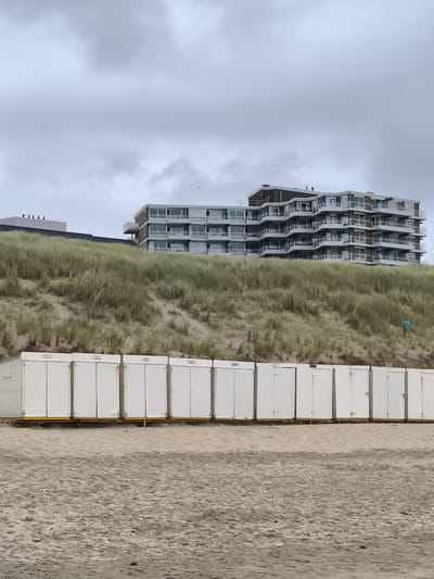 The image depicts a coastal scene with a row of identical white beach huts in the foreground, situated on a sandy beach. Behind the huts, a grassy dune slopes upwards, partially obscuring a multi-story apartment building with numerous balconies and windows. The sky is overcast and grey. The beach huts appear to be uniformly white with dark-colored doors and a subtle yellow trim at their base. The dune is covered in dry, yellowish-green grass. The apartment building is modern in design, with a stepped facade and balconies. The overall environment suggests a slightly chilly or overcast day at the coast. The location is the city of Bergen, Netherlands, specifically its beach area. There are no people visible in the frame.