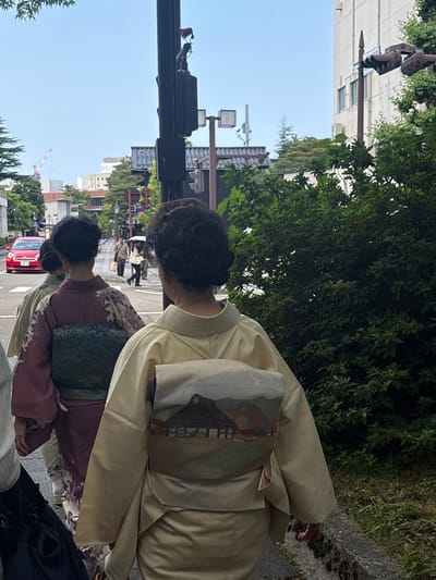 This image captures a daytime street scene in Kanazawa, Japan, under a light blue sky that appears clear or partly cloudy. In the foreground, three women are walking away from the camera, all dressed in traditional Japanese kimonos. The woman closest to the viewer on the right wears a light gold kimono with an obi (sash) featuring a landscape design that includes traditional thatched-roof houses set against mountains. Her dark hair is styled in a neat bun. To her left, another woman wears a deep maroon kimono adorned with a subtle light-colored floral or botanical pattern, complemented by a dark green, textured obi. A third woman, partially visible on the far left, wears a lighter, possibly cream-colored kimono.
In the midground, a red hatchback car, identifiable as a Toyota model, is on the asphalt road, displaying a Japanese license plate with the visible characters 'な 75-76'. On the sidewalk beyond the car and the kimono-clad women, other pedestrians are walking; one man carries a paper bag, and a woman further back holds an open umbrella.
The urban environment features a blend of modern and traditional elements. Multiple utility poles, traffic lights, and streetlights line the road. A prominent black utility pole on the right side of the image supports signal boxes and is topped with a small, light brown animal figurine, possibly a dog or fox. Lush green trees and dense bushes flank both sides of the street, with a significant hedge on the right. In the background, various multi-story buildings are visible, some exhibiting traditional dark tiled roofs, against the light blue sky.