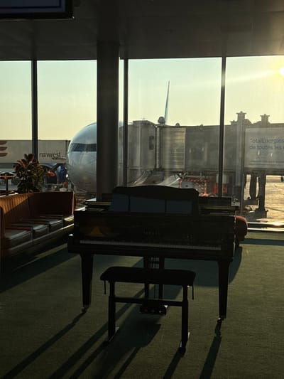 The image depicts an interior airport terminal view in Roissy-en-France, specifically Paris Charles de Gaulle Airport, observed through large windows overlooking an airfield. In the foreground, a dark Yamaha grand piano, with its lid open and a music stand, is positioned centrally on a dark green carpeted floor, accompanied by an adjustable piano bench. The piano and bench cast distinct, elongated shadows across the carpet, indicating a low-angle sun source from the right side of the frame. To the left, brown leatherette sofa seating and a potted plant are partially visible. Beyond the windows, the nose and front fuselage of a large passenger aircraft are visible, parked at a gate with a jet bridge connected to its side. Various ground support vehicles, including one labeled "newrest," are present on the tarmac. A sign displaying "TotalEnergies" in French is discernible on the far right exterior. No individuals are depicted within the frame.