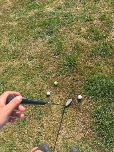 A first-person perspective shows a golf putter poised near three golf balls on a patch of mixed green and brown grass. One ball is white, another white with black spots, and the third yellow and green. A hand grips the putter handle, and parts of a leg and foot are visible in the background. The scene is bathed in bright sunlight, casting shadows from the golf club.