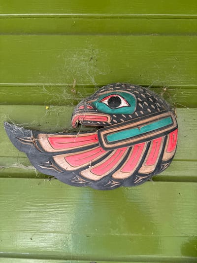 An ornate, stylized avian head carving is affixed to a vibrant green horizontal wooden paneling. The carving displays a black body, a prominent eye with a black pupil, teal iris, and white sclera, a red beak, and layered feathers in red and tan, outlined in black. The entire carving and the surrounding green paneling are extensively covered in fine, white cobwebs and dust. Multiple long-legged spiders, consistent with *Pholcidae* (cellar spiders), are visible within these webs, positioned above and around the carving. The scene is located in Stiperstones, United Kingdom.