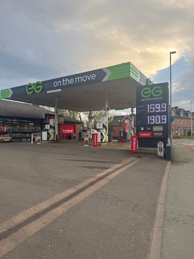 The image shows an EG "on the move" petrol station. The Unleaded fuel price is 159.9 per litre, and Diesel is 190.9 per litre. The station has an Iceland store, Duckhams fuel, and a Costa Express. The location is likely in Welshpool, United Kingdom. The photo appears to be taken from the roadside.