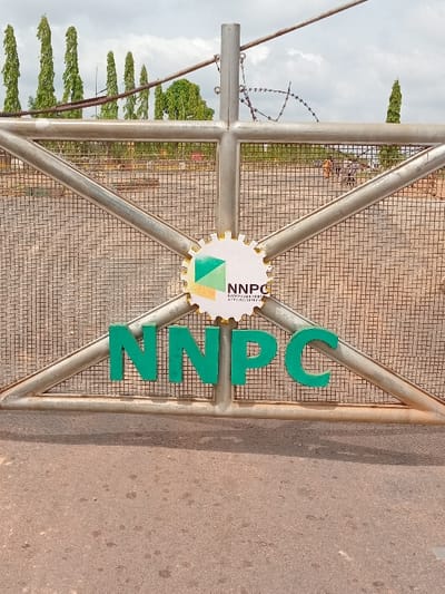 A closed, metal gate with a large green "NNPC" sign on it. The gate is made of metal bars and a wire mesh. It is surrounded by a chain link fence, which has barbed wire at the top. A small, circular, yellow, green, and white logo with "NNPC" and "Nigeria National Petroleum Corporation" written on it, is located above the large green sign. The gate is sitting on a dusty, dirt road. The gate appears to be the entrance to a facility of the Nigeria National Petroleum Corporation. There are trees visible in the background through the wire mesh, which are sparsely populated. The location is a compound in Ungwan Tanko, Kaduna, Nigeria, with coordinates of 10.422 latitude and 7.49 longitude.