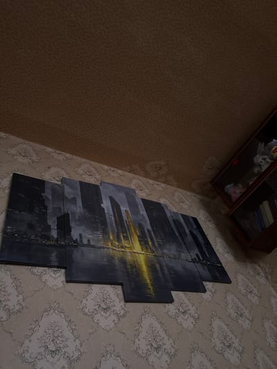 This media file depicts an interior scene, likely a room in a home in Kadiivka, Ukraine. The main subject is a modern, multi-panel abstract artwork hanging on a wall with floral wallpaper. The artwork features a dark, silhouetted cityscape with streaks of golden light, possibly representing city lights at night or a reflection. The wallpaper is a light beige with a repeating pattern of light gray and white floral motifs. To the left of the artwork, a wall covered in textured brown wallpaper is visible, creating a contrast in patterns and colors. In the upper right corner of the image, a portion of a wooden shelf or cabinet is visible, containing several books and some stuffed toys. The lighting suggests an indoor environment, possibly during the day or evening, with artificial light contributing to the overall illumination. There is no discernible activity or event taking place; the scene is static and appears to be a snapshot of a decorated living space. No visible text is present within the artwork or the surrounding environment.