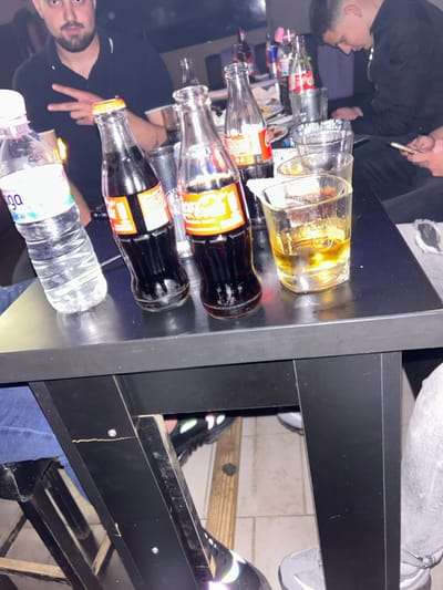 The image depicts an indoor scene within the city of Hvostiane, Bulgaria, likely a bar or restaurant, under dim lighting. In the foreground, a dark rectangular table is laden with multiple beverage containers. From left to right on the table, there is one clear plastic bottle of Devin mineral water, two full glass bottles of Coca-Cola, and a clear glass tumbler containing a golden-amber liquid with ice cubes. In the blurred background, several more glass Coca-Cola bottles, various empty glasses, and other items are visible.

Two males are partially visible seated around the table. On the left, a male with a beard and mustache, wearing a dark polo shirt, looks towards the camera with his left hand raised, forming a "peace" sign. A small orange glow, possibly from a lighter or cigarette, is seen near his hand. On the right, another male with short dark hair, dressed in a dark jacket, is looking down and manipulating a mobile phone with his right hand. Partial views of other individuals' legs and feet, wearing jeans and sneakers, are visible under the table. The floor beneath the table consists of light-colored tiles interspersed with wooden planks.