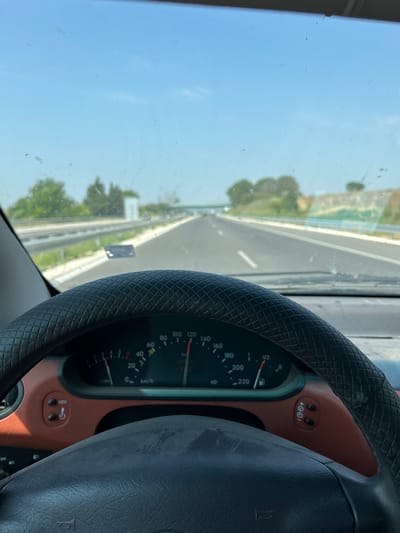 The image shows a first-person view from inside a car, looking forward through the windshield and at the dashboard. The steering wheel, covered in black textured material, is visible on the right side of the frame. To the left of the steering wheel, the car's dashboard displays a speedometer with markings up to 220 km/h and a fuel gauge. Above the speedometer, there are two buttons with icons that appear to be related to cruise control or vehicle settings.
The view through the windshield reveals a highway stretching into the distance. The road is paved, with lanes clearly marked. On either side of the highway, there are grassy verges and some greenery, suggesting a rural or suburban setting. The sky is a clear, bright blue, indicating a sunny day. The angle of the sun suggests it is likely daytime, perhaps late morning or afternoon. There are no people visible in the image. The overall scene conveys a sense of travel on an open road under pleasant weather conditions.
The image includes visible text on the dashboard instruments: "100", "120", "80", "140", "60", "160", "40", "180", "1/2", "20", "200", "0", "km/h", "220", "0000", and "+00".