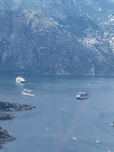 This aerial shot captures a coastal area, likely the Bay of Kotor in Mirac, Montenegro. The image shows rocky mountains on the left side, adjacent to the bay's blue waters. Multiple cruise ships are sailing in the bay, along with other smaller boats, evidenced by their wakes. The right side features small rock islands, possibly with settlements. The environment is bright, indicating daylight.
