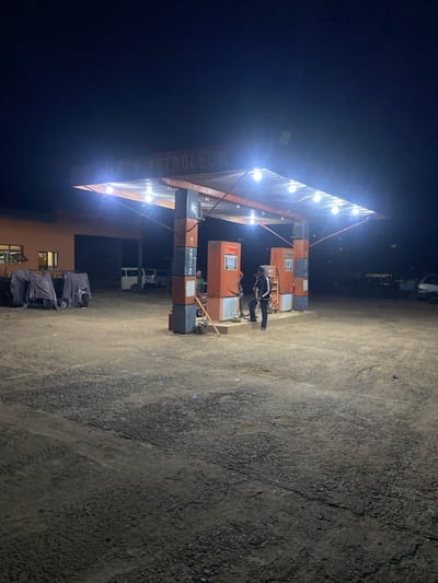 This image depicts a nighttime scene at a gas station identified as HACO PETROLEUM in Sasaro, Nigeria. The gas station features a central canopy illuminated by multiple bright white light bulbs attached to its underside. Four fuel pumps, primarily orange and gray, are positioned beneath the canopy. One supporting pillar explicitly displays "NO SMOKING" vertically in white lettering. The ground beneath and surrounding the station is unpaved, consisting of dark gravel and dirt.
Three individuals are present under the canopy. Two males are visible near the central fuel pumps; one wears a dark jacket and trousers, standing and leaning slightly, while the second is partially obscured behind him. A third male, wearing a green shirt, is partially visible further to the right near another pump.
To the left of the gas station structure, a building with an orange-brown facade and two visible windows is present. In front of this building, several large objects are covered with dark tarps or sheets. In the background, a white van and another dark-colored vehicle are parked on the left-center, and a light-colored car is partially visible on the far right. Numerous small insects are visible in the air, attracted to the bright lights under the canopy. The sky is dark, indicating night conditions.