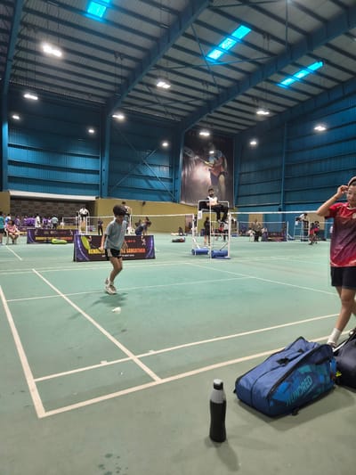 The image depicts an indoor badminton court located in Phagwara, India. The court is illuminated by overhead lights and features a green synthetic floor with white court lines. Several individuals are present on and around the court, participating in or observing a badminton match. In the foreground, a young boy in a grey t-shirt and black shorts is mid-jump, about to hit a shuttlecock. To the right, a person wearing a red and black t-shirt and black shorts is partially visible, with their hand touching their head. Two large blue badminton bags and a black water bottle are placed on the court floor near the foreground. In the background, there are spectators, officials seated on elevated chairs, and other players. A large banner featuring an image of a badminton player hangs on the back wall, along with tournament advertisements. The overall environment suggests an active sporting event taking place within a modern sports facility.