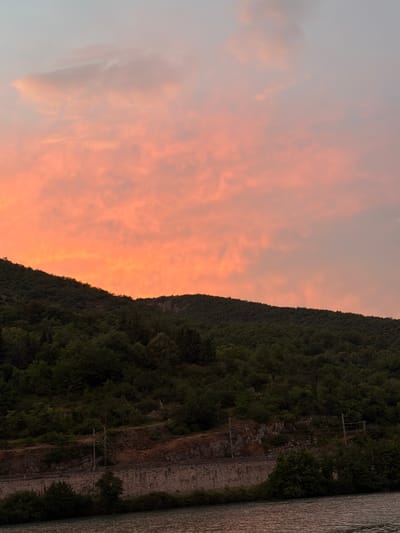 The image depicts a scenic view of the Rhône River and a forested hillside at sunset in Loriol-sur-Drôme, France. The sky is filled with vibrant orange and pink clouds, characteristic of a sunset. The hillside is densely covered with dark green trees, with a stone wall and railway tracks visible along its base. The river in the foreground reflects the colors of the sky. There are no people or animals present in the image. The overall scene conveys a sense of natural beauty and tranquility.