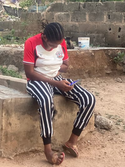 An outdoor scene in Idiroko, Nigeria, features a female individual seated on a concrete ledge. She is attired in a red and white short-sleeved top with a visible logo, black and white vertically striped jogger pants with elastic cuffs, and mismatched flip-flops (one red-soled, one brown-soled). Her hair is styled in tight braids. The individual holds a smartphone with both hands, operating it; the screen emits a blue illumination. The ground consists of bare earth with scattered rocks and debris. In the mid-background, a concrete block wall is present, upon which rests a white plastic bucket displaying the words "Blue Paints." A small, dark-colored goat is situated behind the bucket, near the wall. Sparse vegetation and distant rudimentary structures are visible in the background.