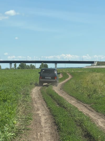 The image shows a black Niva SUV driving on a dirt road through a grassy field. In the background, a large bridge spans across the horizon. The sky is blue with a few white clouds. The scene appears to be in a rural area, possibly near Tarasovo, Russia, given the context. The car is kicking up dust as it moves along the road. The vegetation is green and lush, suggesting a warm, possibly summer, day. The overall impression is one of a journey through the countryside.