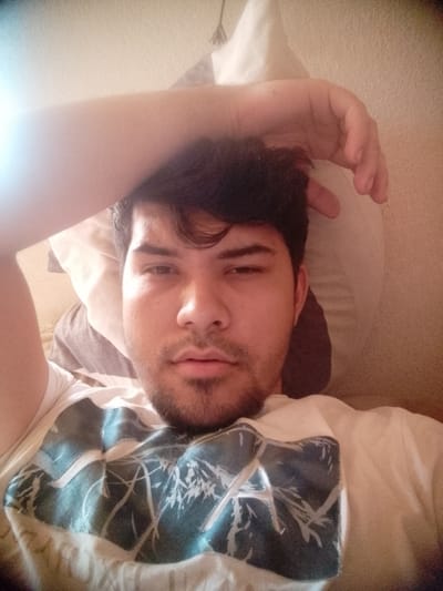 A person is lying down, possibly on a bed, with a pillow behind their head. They are wearing a white t-shirt with a blue and white abstract pattern. Their arm is raised and resting on their forehead. The background is a light beige or white wall. The location is unknown, but based on the user instructions, the location would be in Osenets, Bulgaria.