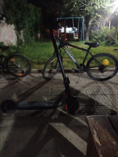 The image shows a scooter and two bicycles parked on a paved surface. Behind them is a grassy area with trees and a swing set in the background. The location appears to be Gavrailovo, Bulgaria. The scene is dimly lit, suggesting it is nighttime. The focus is on the objects and setting, with no people visible.