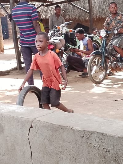 A young boy, wearing an orange t-shirt and dark shorts, runs with a tire in an outdoor, dirt-ground setting. He is on the right side of the frame, his body turned to the left and his head looking towards the left as well. His right hand is extended towards the tire, and his left arm is bent at the elbow. The tire is a dark, worn-out rubber with a visible tread pattern.
In the background, several people and motorcycles are visible. A man in a striped blue and red shirt stands to the left, his back to the camera. Behind him, another person in a yellow shirt is partially visible. Further back, two young men are seated on or near motorcycles. One is wearing a patterned shirt and appears to be on a motorcycle, while another is wearing a blue tank top and dark pants, looking down at something in his hands. Multiple motorcycles, some with visible handlebars and wheels, are parked in the area, suggesting a gathering or marketplace. The scene appears to be under a thatched roof structure, providing shade. The lighting suggests it is daytime. The ground is dusty and uneven. The overall atmosphere seems casual and active, possibly indicating a playful moment or a functional task involving the tire. There is a cracked concrete structure in the foreground that extends across the bottom of the image.