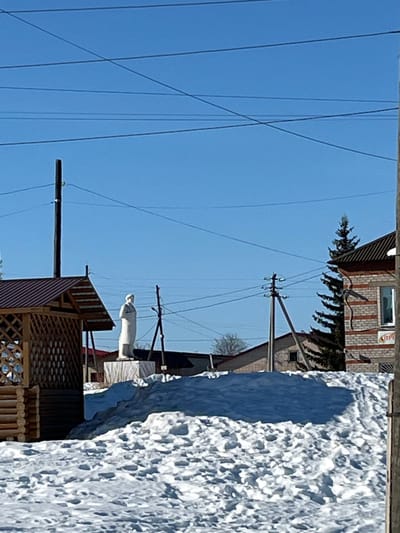 This image depicts an outdoor scene in the city of Сундур, Russia, characterized by a bright blue sky and snow-covered ground. In the mid-ground, a white full-body statue of a male figure stands erect on a light-colored rectangular plinth, which is situated on a raised mound of snow. The figure wears a long coat or robe and faces towards the left of the frame. To the left of the statue, a wooden gazebo with a dark brown sloped roof and latticework sides is partially visible, with a log-like base. Numerous utility poles (both wooden and metal) and overhead power lines traverse the sky. In the background, on the right, a two-story brick building with alternating light and dark horizontal banding is visible, featuring a window on the upper floor and a dark roof. A tall, dark green evergreen tree stands adjacent to this building. Further in the background, other structures with dark roofs and lighter walls are partially discernible. The ground in the foreground is entirely covered with irregular patches and mounds of snow. No active people or specific actions are observed.