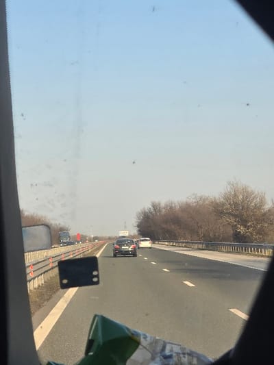 The scene depicts a multi-lane asphalt highway, likely in Trilistnik, Bulgaria, viewed from inside a moving vehicle through a moderately soiled windshield. The sky is clear and pale blue.
In the foreground, the bottom edge of the image reveals part of the interior of the capturing vehicle, including a green and white object (possibly packaging material) and a dark, rectangular device, possibly a dash camera or mounting bracket, affixed to the windshield. A portion of a vehicle's side mirror or its housing is visible on the far left.
The highway extends into the distance, featuring two visible lanes of travel in the same direction, separated by broken white lines. A solid white line delineates the right shoulder. Metal guardrails run along both sides of the highway, with orange safety markers or reflectors visible on the left guardrail.
Several vehicles are present on the highway. A dark-colored sedan is positioned in the left lane, followed by a white sedan or hatchback further ahead. Another white vehicle is partially visible on the right shoulder or in the slow lane further down the road. In the far distance, two large trucks, one with a red cab and another with a dark cab, are visible on the left side of the highway. A blue highway sign on an overhead gantry structure is visible above the left lane in the middle distance; its text is illegible.
The environment adjacent to the highway consists of bare, deciduous trees and brush, suggesting a winter or early spring season. No individuals are distinctly visible.