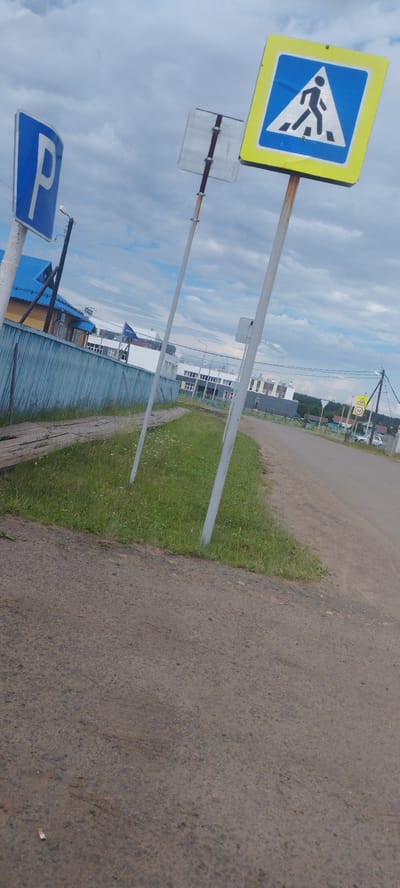 The image shows a street scene with several traffic signs in Pokosnoe, Russia. In the foreground, a dirt road curves to the right, with a strip of green grass and some wooden planks bordering it on the left. Two traffic signs are prominently visible: a blue square sign with a white letter "P" (indicating parking) is on the left, and a yellow square sign with a black pedestrian symbol inside a blue triangle (indicating a pedestrian crossing) is on the right. Both signs are mounted on metal poles. In the background, a light blue wooden fence runs along the left side of the road, with a yellow building with a blue roof behind it. Further back, several multi-story buildings with various colored facades are visible, along with trees and a cloudy sky. Another yellow triangular sign with a number "20" (likely a speed limit sign) is visible on the right side of the road. The overall scene depicts a relatively quiet, semi-urban or rural environment.