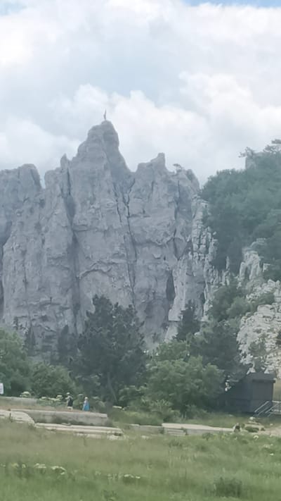The image depicts a mountainous landscape with rocky formations and dense vegetation. A prominent peak in the center of the image has a solitary figure standing atop it, appearing as a silhouette against the sky. Below the rocky cliffs, there is a treeline with various shades of green foliage. In the foreground, there is a grassy area with some people scattered around, some standing and some walking. There is also a structure with a dark roof and stairs leading up to it on the right side of the image. The sky is partly cloudy with a bright overcast. The scene appears to be a natural recreational area, possibly a tourist destination given the presence of people and the elevated viewpoint. The location is identified as Okhotnichye, Ukraine.