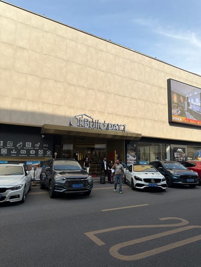 The image shows the exterior of a building in the Chancheng District, China, possibly a shopping center or showroom. The building's façade is tan and features a large sign reading "OkBuildDAY1." A screen advertising "20000m² One-Stop Shop" is displayed above the entrance. Parked in front of the building are several cars, including a red SUV, a dark-colored SUV, a white Mercedes, and another dark SUV. There are a few individuals standing near the entrance. A number "20" is painted on the asphalt in large numbers. Several icons are present below the entrance. The icons contain images of doors, windows, sanitary ware, furniture, lighting, and more. License plates from China are visible on the cars.