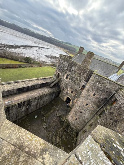 An aerial-angled photograph, taken from a high vantage point within Blackness Castle, Blackness, United Kingdom, shows the castle's stone architecture and surrounding landscape under an overcast sky. In the immediate foreground, a weathered stone parapet covered in white lichen provides the viewing platform. Below, the castle's internal structure reveals deep, rough-hewn stone walls forming a central, irregular courtyard filled with dark, rocky terrain. A single individual, dressed in dark attire, stands near a shadowed arched entrance at the base of a prominent stone wall on the right side of the courtyard. An elevated stone walkway, equipped with black metal railings, traverses the middle distance, leading to a set of bare wooden steps that ascend towards another entrance in the castle wall. Beyond the castle walls, a strip of vibrant green grass is visible, followed by an expansive, pale landscape featuring what appears to be a snow-dusted or ice-covered body of water or mudflats, flanked by tree lines and undulating terrain with patches of snow and bare fields. Several stone chimney stacks rise from the roofs of the castle buildings, visible in the upper right.