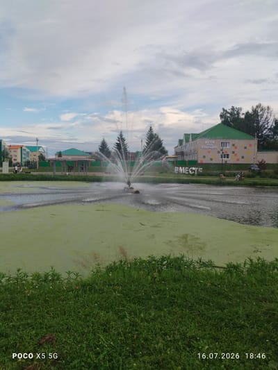 The image shows a public park in Aksubayevo, Russia, on a cloudy day. In the foreground, lush green grass lines the edge of a pond covered in a thick layer of green algae. A fountain sprays water in the center of the pond, creating ripples. In the background, buildings with green roofs are visible, one of which has colorful square tiles and the Russian text "ДЕТИ НАШЕ БУДУЩЕЕ!" (Children are our future!). Large, three-dimensional letters spelling "ВМЕСТЕ" (Together) stand on the grassy bank near the pond. Several people are present in the scene, including individuals walking near a crosswalk, sitting on a bench under a gazebo, and two people seated on another bench in the middle ground. A car is parked near one of the buildings. The timestamp in the lower right corner indicates the photo was taken on July 16, 2026, at 18:46. The camera information "POCO X5 5G" is displayed in the lower left corner.