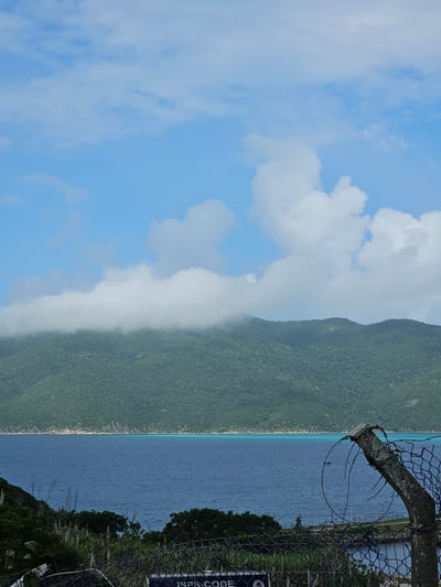 This image depicts a coastal landscape in Arraial do Cabo, Brazil. The scene is dominated by a light blue sky with scattered white cumulus clouds, with some larger formations present on the right. A large, verdant mountain range covered in dense green vegetation stretches across the background, with its peaks on the left partially obscured by a horizontal band of white fog or low-lying clouds. In the middle ground, a vast expanse of deep blue water, likely the ocean or a bay, is visible, featuring a distinct narrow strip of turquoise water where it meets the distant, vegetated shoreline.
The foreground contains green foliage, including several cactus-like plants on the left. A dark-colored chain-link fence extends across the lower portion of the frame, partially obstructing the view. On the right side of the fence, a weathered, gray concrete post is visible, topped with rusted, tangled barbed wire. A dark blue sign with white lettering, partially obscured by the fence at the bottom center, displays the text "ISPS-CODE." A small, dark-hulled boat with a light-colored cabin is visible on the water in the middle distance, positioned to the left of the concrete fence post. No human figures are explicitly visible in the scene.