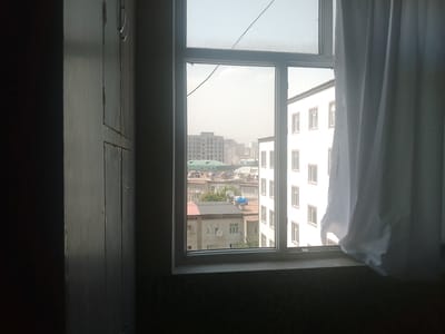 The image shows a view through a window in Vahdat, Tajikistan. The window is partially obscured by a white curtain on the right side. Outside, a cityscape is visible, with multiple buildings of varying heights and architectural styles. Some buildings appear to be residential apartments, while others might be office buildings or public structures. The sky is hazy, suggesting either pollution or a dusty atmosphere common in the region. In the foreground, on the left, part of a dark, wooden structure, possibly a door or cabinet, is visible. The overall scene conveys a sense of an urban environment under natural light, likely during daytime.
