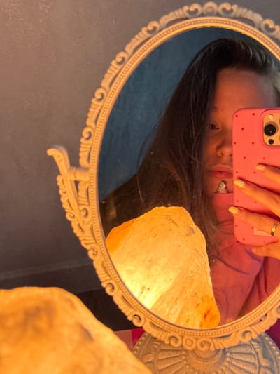 The image is a selfie taken by a young person reflected in an oval mirror with an ornate, decorative frame. The person is holding a bright pink phone with small red polka dots, which is partially obscuring their face. They are wearing a pink top and have yellow nail polish with a speckled design. A ring is visible on their right ring finger. To the left of the mirror, a large, rough-hewn, illuminated object, likely a salt lamp, glows with an orange-yellow light. The background consists of a dark, textured wall. The scene appears to be indoors, possibly in a bedroom or dressing area, and is lit by the warm glow of the lamp. The context of Tliustenkhabl', Russia is not directly evident from the visual information in the image.