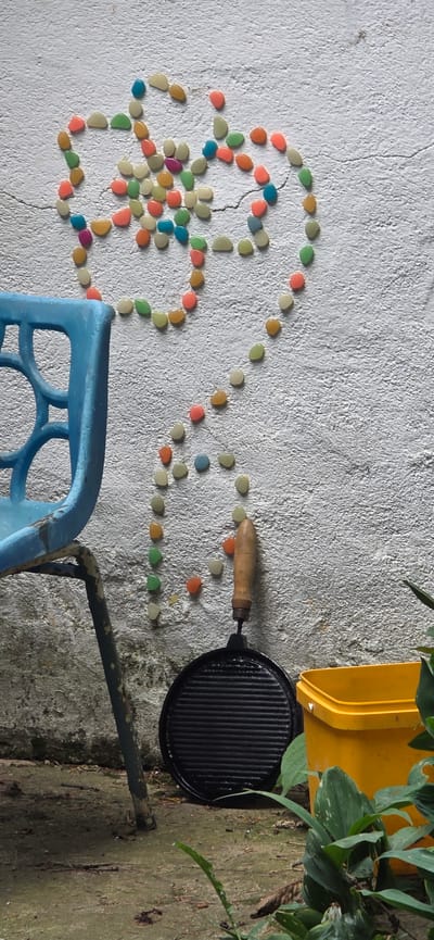 This vertical shot captures an outdoor scene against a textured white wall in the city of Partizanin, Bulgaria. Dominating the upper two-thirds of the wall is an arrangement of numerous small, colorful, smooth pebbles, forming a stylized four-petal flower or clover-like shape at the top, with a wavy, stem-like trail descending towards the bottom center. The pebbles are varied in color, including shades of light green, orange, pink, blue, yellow, and beige. The white wall exhibits a rough, stucco-like texture with visible cracks, notably a horizontal one running through the pebble arrangement's upper portion.

In the lower left quadrant, a portion of a faded blue plastic chair is visible, showing part of its backrest with circular cut-outs. The chair's metal frame, painted a dark green but heavily rusted and chipped, extends towards the bottom left. Directly below the pebble trail and resting on the ground against the wall is a black, circular cast-iron griddle or pan with a ridged cooking surface and a short, light brown wooden handle pointing upwards. To the right of the griddle, a bright yellow plastic bucket or container is partially visible, obscured by lush green foliage emerging from the bottom right corner of the frame. The ground beneath the objects appears to be concrete or paved, showing some dirt, debris, and possibly moss.