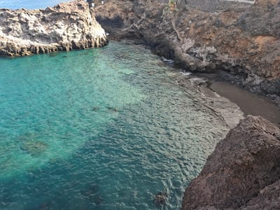 This is an aerial view of a cove in Santiago del Teide, Spain. The water is a clear, vibrant turquoise and laps against a dark sand beach and rocky cliffs. The cliffs are rugged and uneven, with some vegetation visible on the upper ledges. There are no people or animals in the frame. The scene depicts a natural coastal landscape.