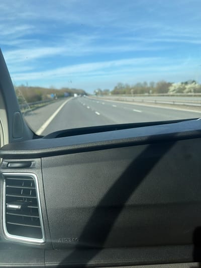 The image displays a forward-facing view from inside a vehicle, positioned on a multi-lane highway in Chieveley, United Kingdom. The lower portion of the frame shows a dark, textured vehicle dashboard, featuring a silver-trimmed air vent on the left and an embossed "AIRBAG" label below it. A distinct diagonal shadow is cast across the dashboard from the upper right. Through the windshield, an asphalt highway stretches into the distance, marked by solid and broken white lines indicating multiple lanes of travel. At least three lanes are visible in the immediate direction of travel. The highway features a central reservation with a metal crash barrier. Trees, some appearing dormant, and scrub brush line the highway on both sides, with some white flowering vegetation visible on the right. Several vehicles are distantly visible on the highway. The sky is bright blue with scattered thin white clouds, consistent with clear daytime conditions.