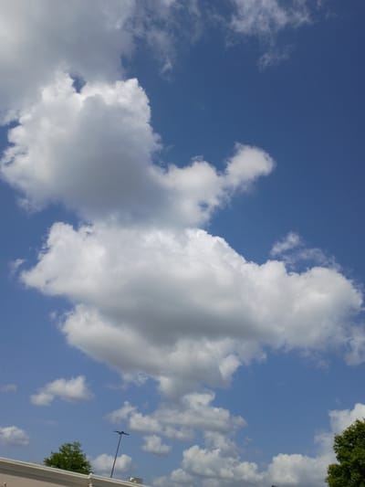The image shows a cloudy blue sky with a few trees and a light pole visible in the lower portion. The clouds are white and fluffy, with some appearing larger and more dense than others. The sky is a clear, bright blue. A single light pole with a dark fixture at the top stands in the mid-ground, and a lush green tree is to its left. The lower edge of the image shows a white building with a slight overhang. The scene appears to be outdoors, possibly in a park or suburban area.