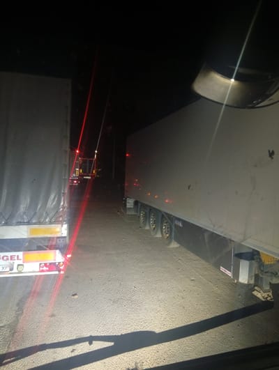 At night, several semi-trailer trucks are parked on a dirt road. The camera is positioned from within a vehicle, looking forward. The closest truck, on the left, is gray with a reflective white and yellow stripe on the rear. The second truck from the left, further down the road, is dark with red taillights and yellow lights on the roof. The truck on the right is also gray, with its side visible, showing multiple wheels and a dark mudguard. The ground is a rough, gravelly dirt surface. The environment is dark and appears to be rural, with trees visible in the background. The location is specified as Mamalyha, Ukraine.