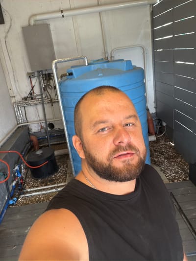A man with a beard, wearing a black t-shirt, poses for a selfie in an outdoor utility area. Behind him, a large blue water tank stands prominently, connected to various pipes and plumbing fixtures. To the left, a grey electrical panel and insulated pipes are mounted on a white wall. The ground is covered with gravel. A slatted grey fence is visible on the right side of the frame. The image appears to be taken in Sergey-Pole, Russia, likely a residential or industrial setting.